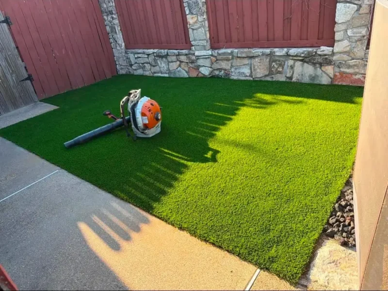 Artificial turf courtyard beside a stone and wood fence, with cleanup equipment