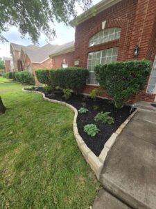 Curved autumn tan stone flowerbed border with fresh dark mulch, new plants and trimmed hedges beside a brick home